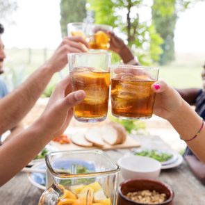 Glasses filled with iced tea clink together, surrounded by smiling people at an outdoor table with food.
