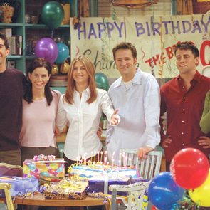 Six people smile at a birthday party, standing in front of a cake with candles. Colorful balloons and a 