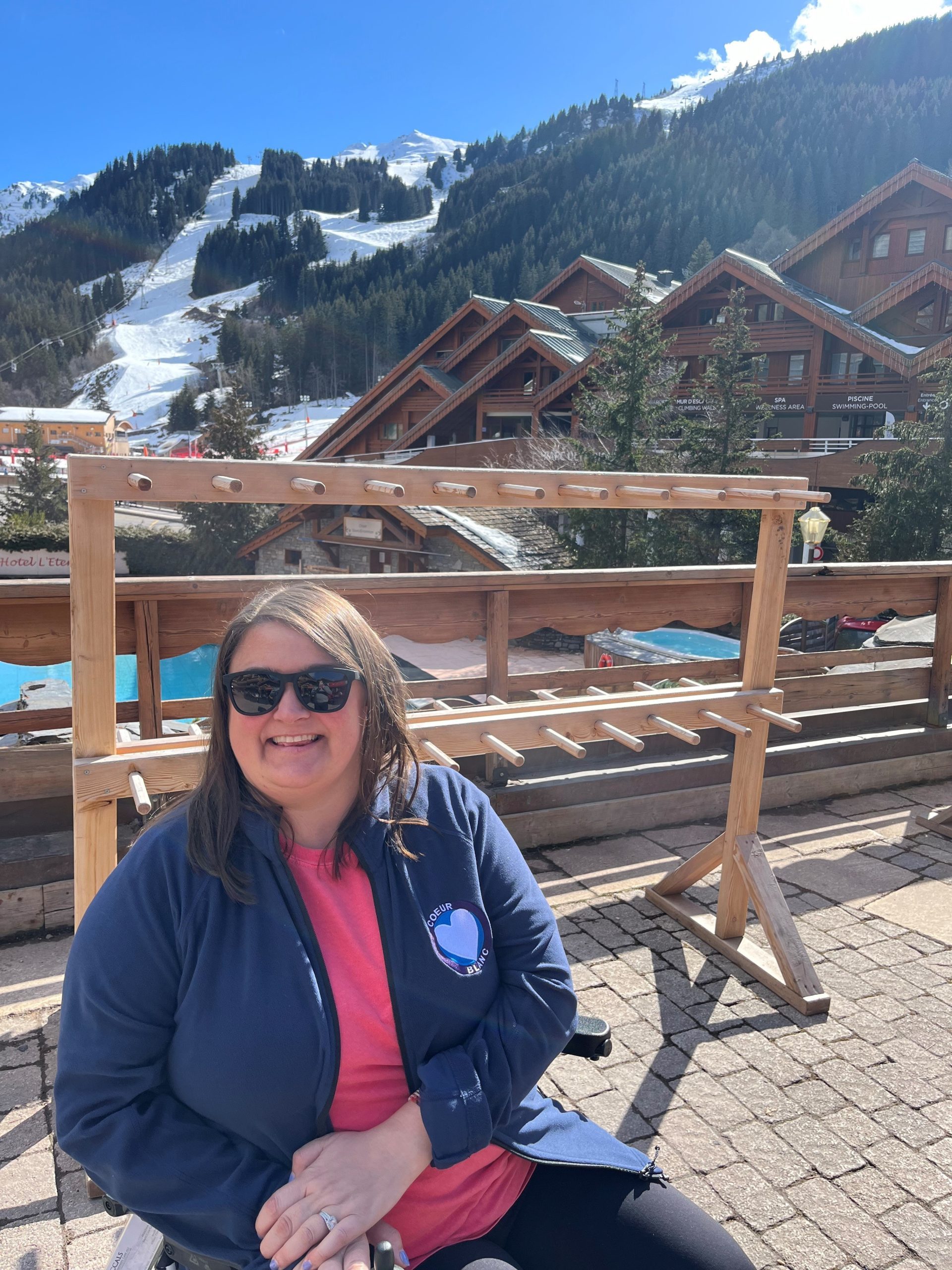 A smiling person sits outdoors wearing sunglasses. Behind, wooden racks and alpine chalets provide a scenic snowy mountain backdrop under a clear blue sky.