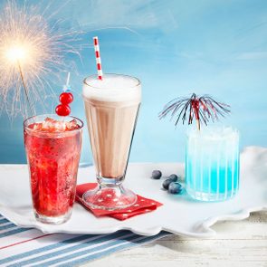 Three colorful drinks sit decorated on a white tray; one has a sparkler, another has a red straw, and the third a cocktail umbrella.