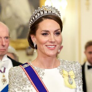 A woman in a jeweled tiara smiles, wearing a pearl necklace and formal attire, surrounded by elegantly dressed guests in a luxurious setting.