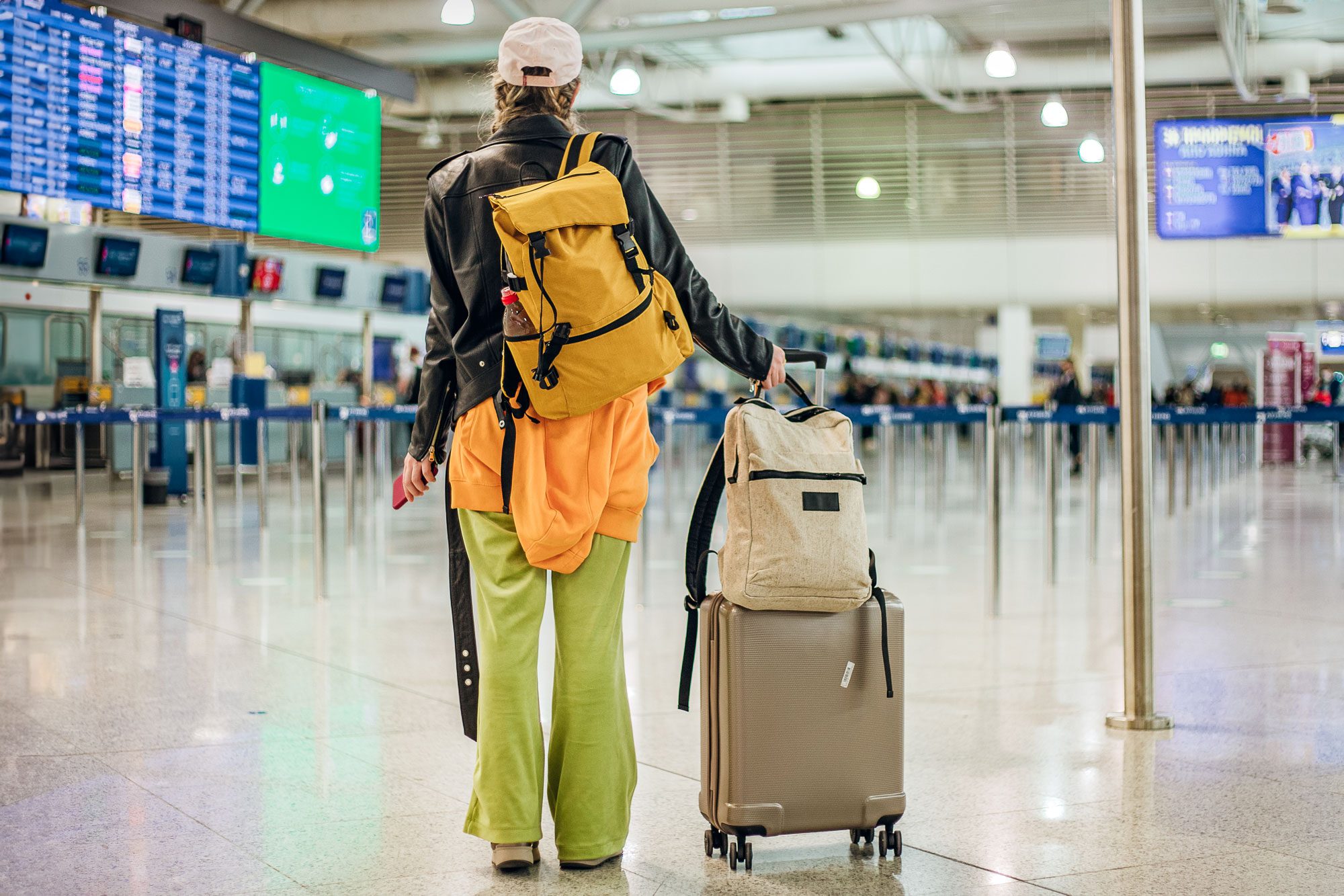 Traveler pulling suitcase, wearing a yellow backpack and bright clothing, walks through an airport terminal with flight information displays overhead.