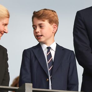 A boy talks to a woman, both in formal suits, standing near a man, likely at an event.
