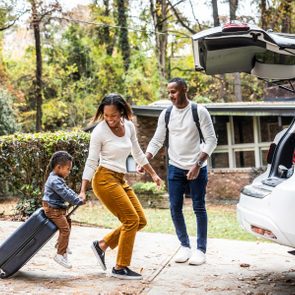 A family packs a white SUV for a trip; a child rides a suitcase, smiling, amidst autumn trees and a house.