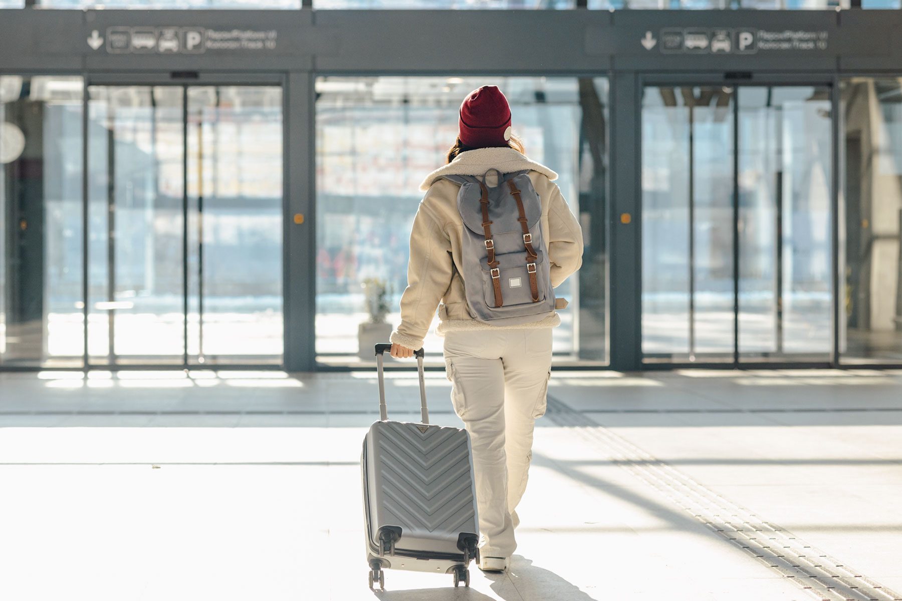 Traveler pulls suitcase, wearing backpack and winter clothing, walking through bright, modern airport terminal.