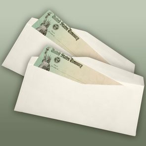 Envelopes containing U.S. Treasury checks are partially open, displaying a Statue of Liberty graphic, against a plain green background.