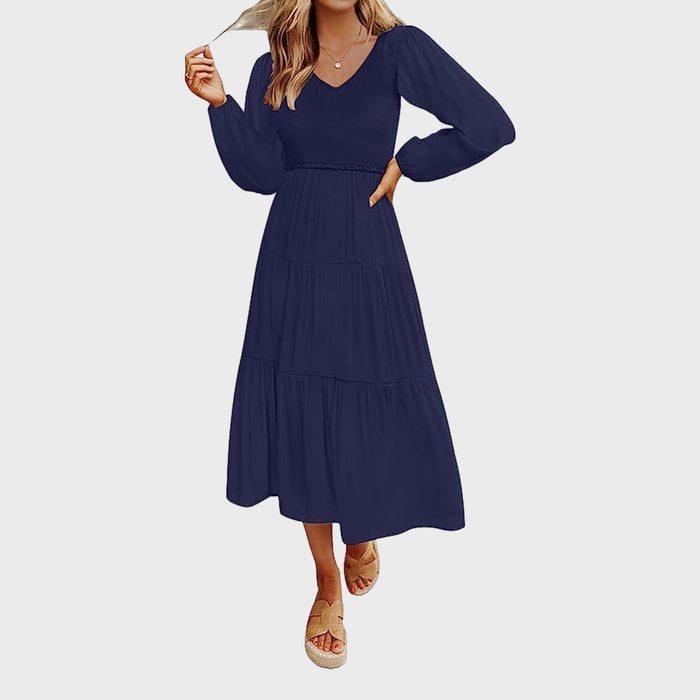A woman stands wearing a long-sleeved, navy blue dress with a V-neck, holding her hair, against a plain white background.