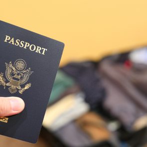 Passport held by a hand, showing the cover design, with a blurred, open suitcase in the background on a beige carpet.