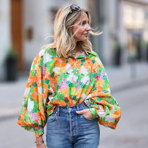 A person wearing a colorful, floral blouse and jeans walks along a city street, with sunglasses resting on their head.