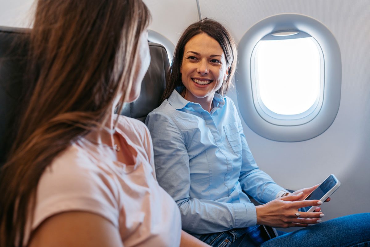 Two Young Female Friends Using Phone And Talking In An Airplane