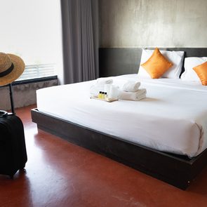 A modern hotel room with a neatly made double bed featuring white linens and two orange pillows. A black suitcase and a wide-brimmed straw hat are near the bed. The room has a large window with a view of greenery outside.