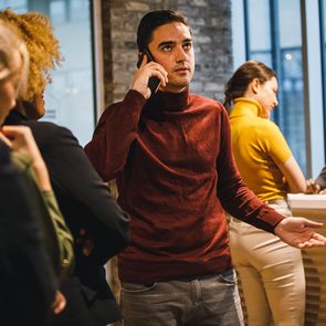 Man in line to check in at a hotel holds a phone up to his ear with an exasperated expression