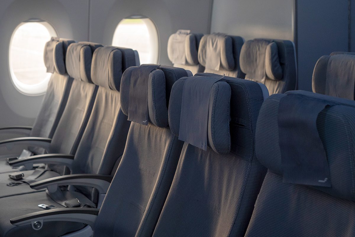 Rows of empty airplane seats with dark gray upholstery and headrests, next to windows letting in natural light.