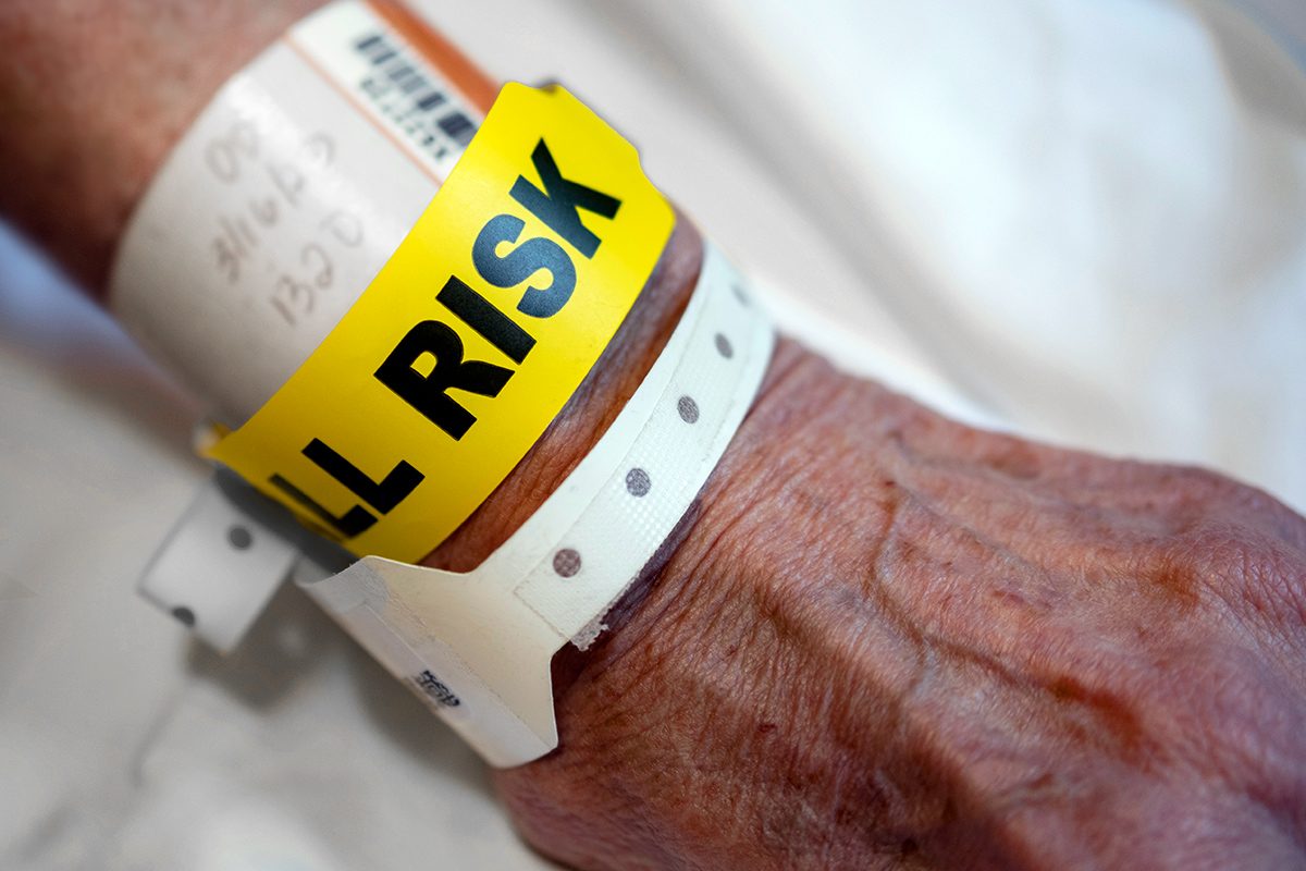 Close-up of an elderly person's wrist wearing hospital identification bands, including a prominent yellow band labeled "FALL RISK," indicating a medical alert for increased fall risk.