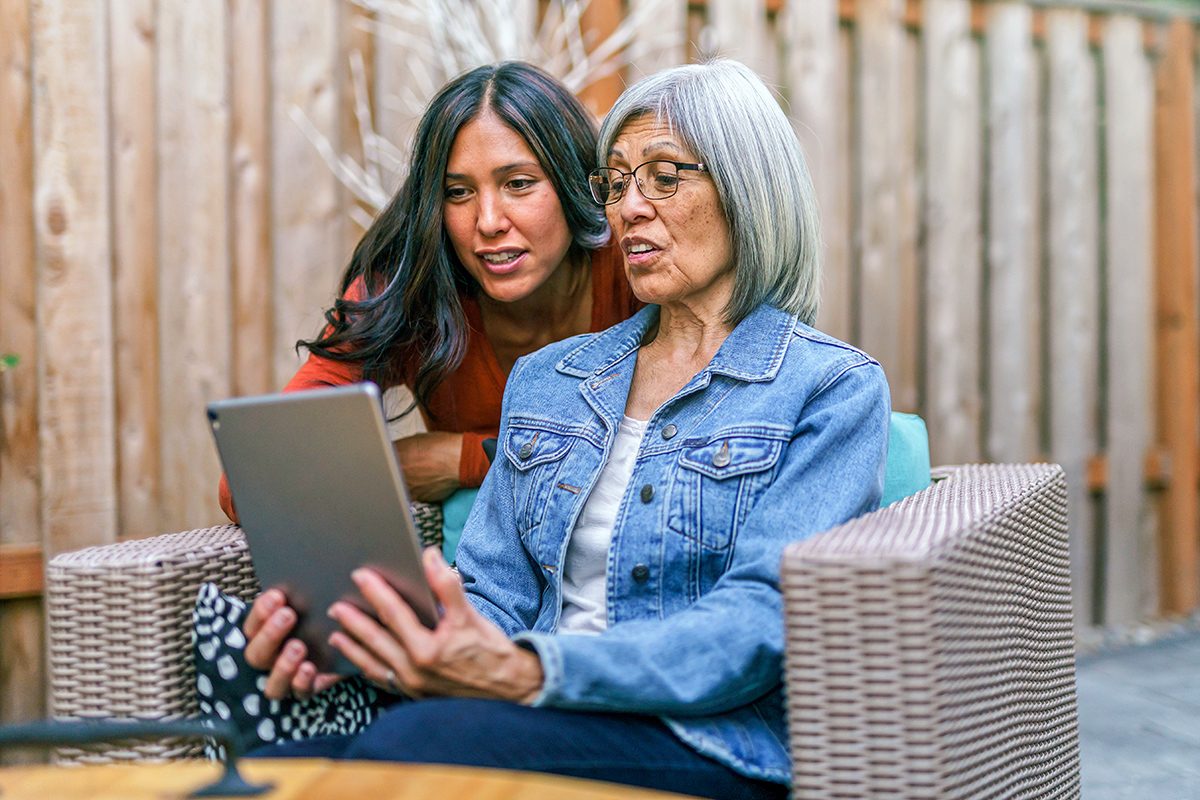Two women, one older and one younger, sit together outdoors on a patio. They are looking at a tablet screen, appearing engaged and focused, with a wooden fence in the background.