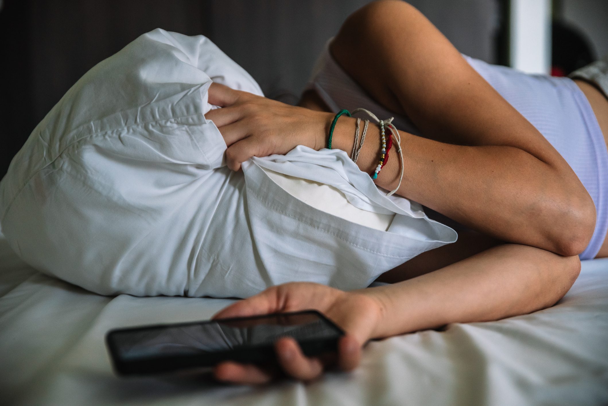 Young woman lying on a bed covering her face with a pillow and holding her cell phone in her hand.
