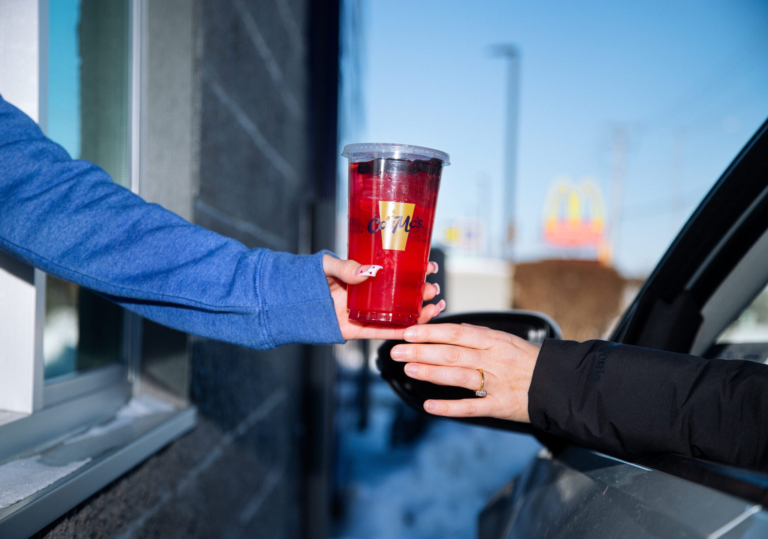 Arm from drive thru hands a CosMc's red drink to an arm reaching from a car