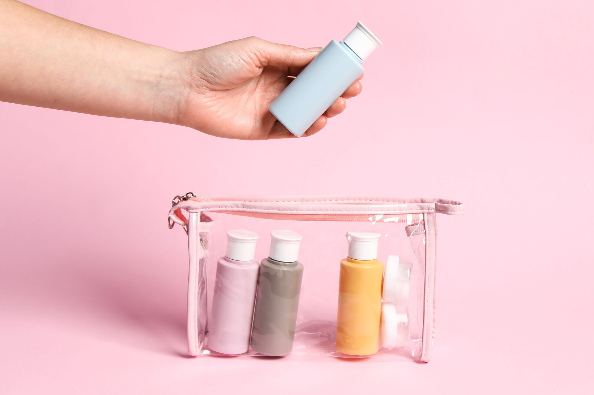 Cosmetic travel kit. Woman putting small bottle with personal care product into plastic bag against pink background, closeup