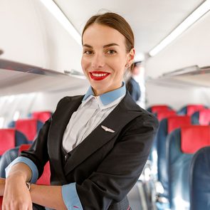 Cheerful woman stewardess or air hostess looking at camera and smiling while placing hand on passenger chair in aircraft