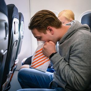 A young man is sitting on an airplane, being sick into a sick bag.