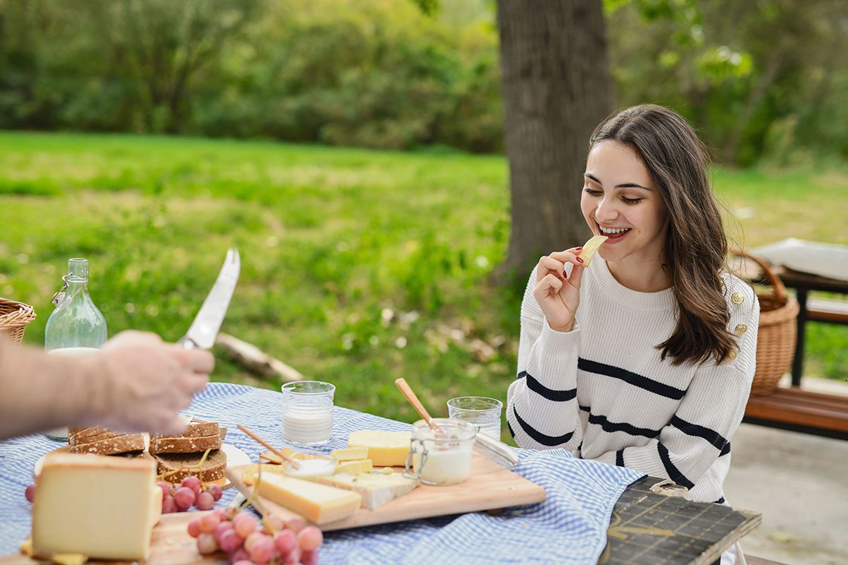 Woman Sitting at Picnic Table Tasting Piece of Freshly Sliced Cheese Enjoying