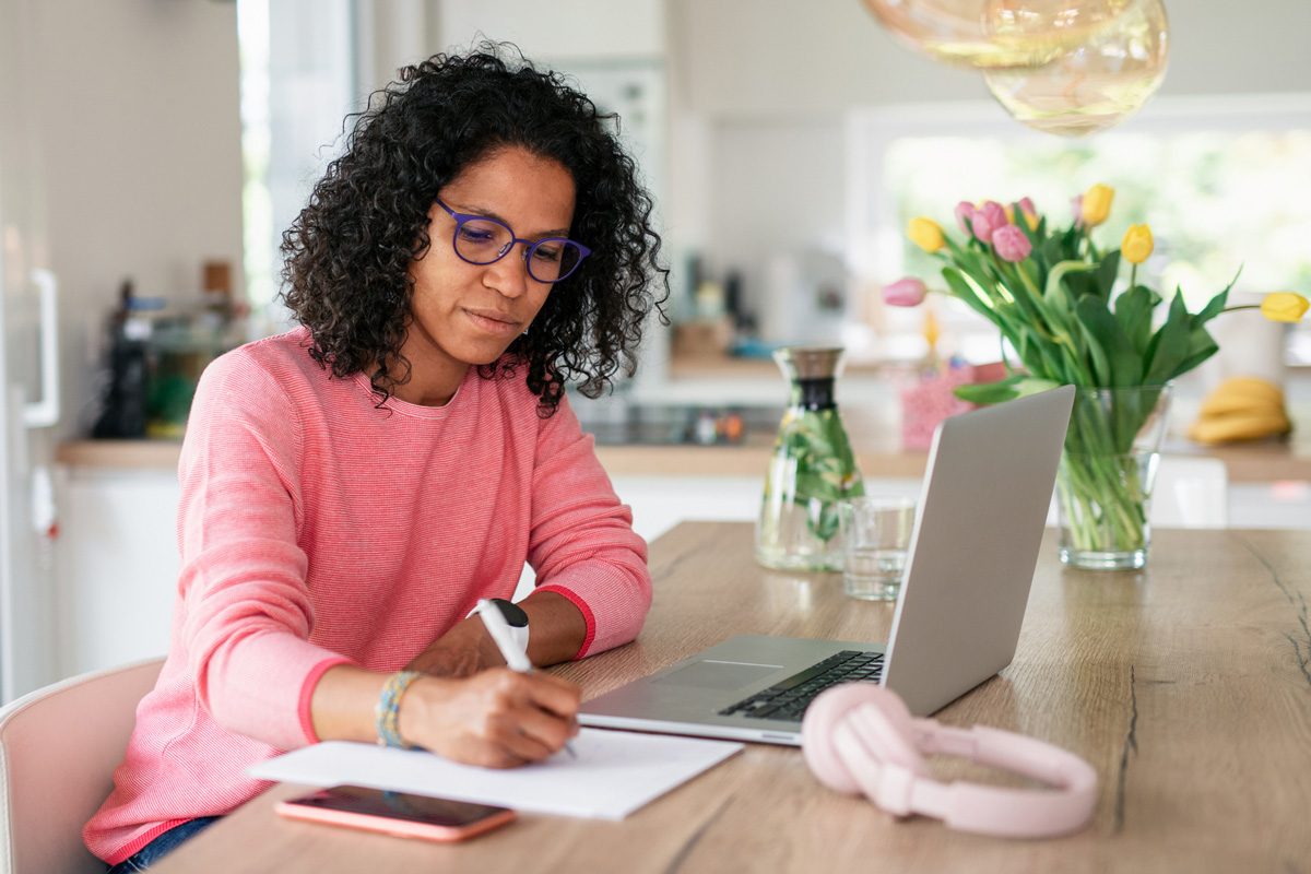 woman writing in notepad while using laptop at home