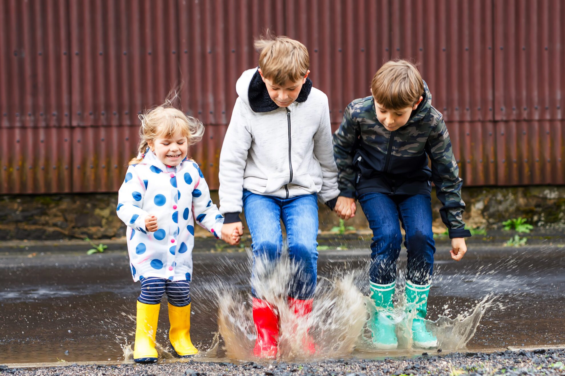Three children, toddler girl and two kids boys wearing red, yellow and green rain boots and walking during sleet. Happy siblings jumping into puddle. Having fun outdoors, active family