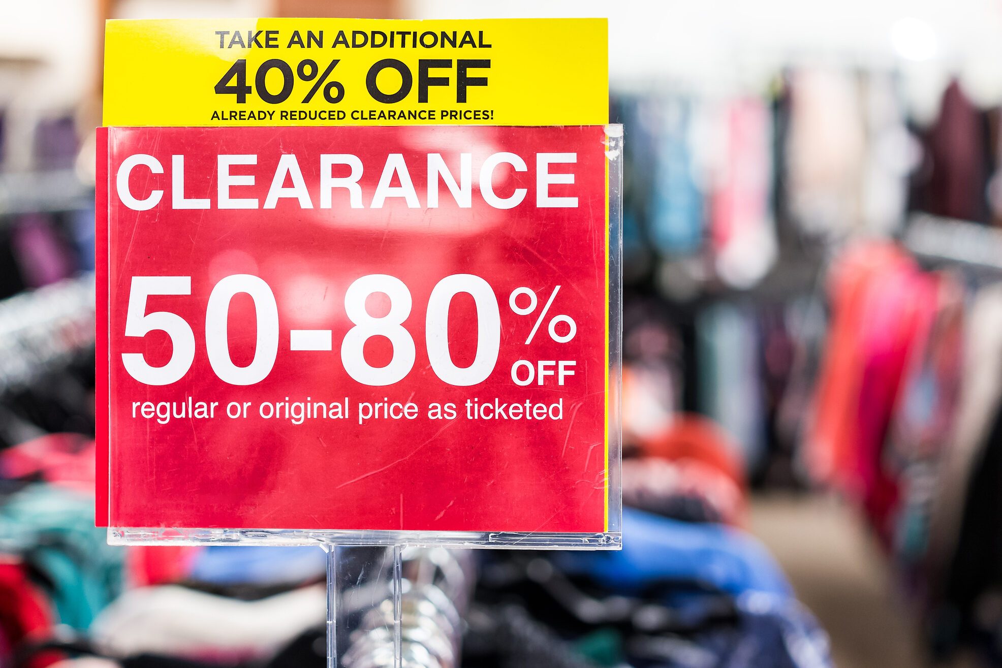 Many clothes bokeh in discount store, garments hanging on racks hangers row closeup with large red clearance sign, reduced prices