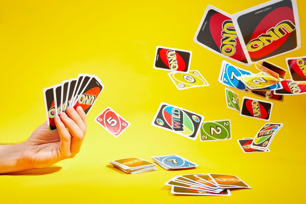 A hand holds up a fan of cards on the left. On the right, a fan of cards is face down and a slew of cards are falling in the air; yellow background