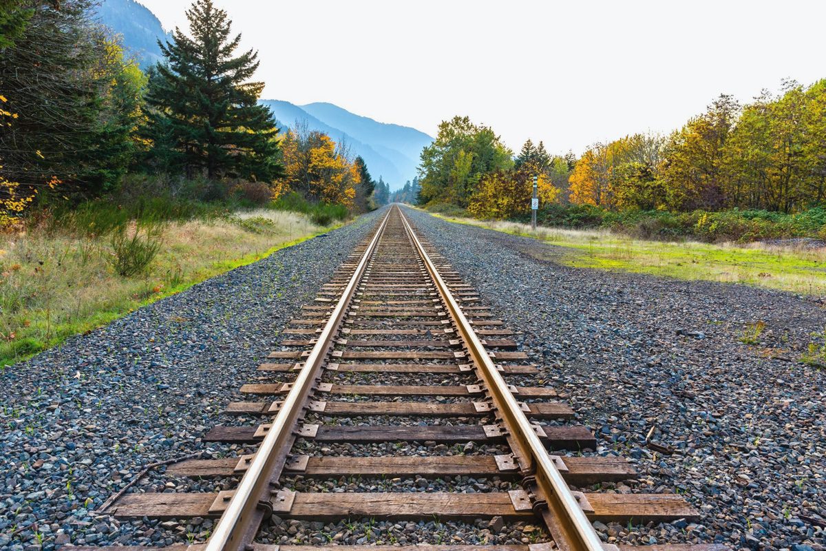 Railway tracks stretch into the distance, flanked by grassy fields and vibrant autumn trees, with mountains visible in the background.