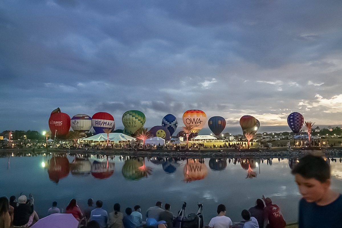 A row of colorful hot air balloons is lined up along the edge of a calm lake at dusk, reflecting in the water, with many people gathered on the shore watching the event under a cloudy sky.