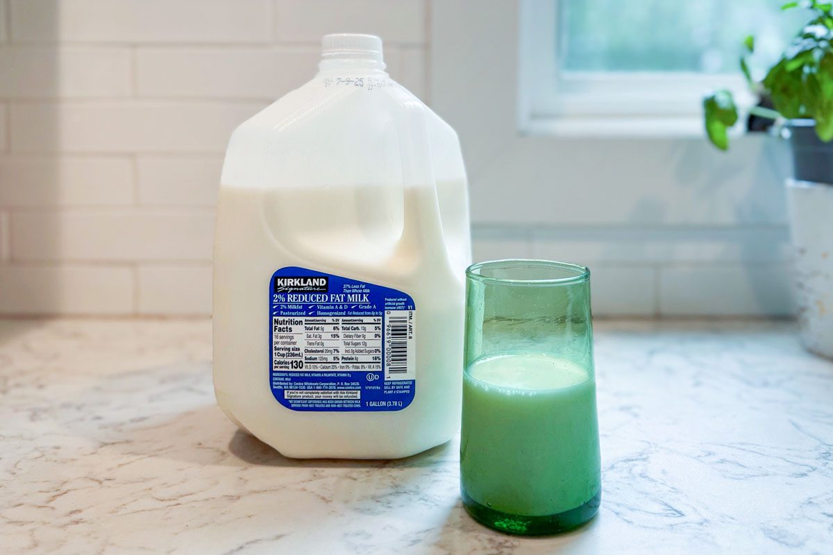 Gallon of Costco's Kirkland Signature 2% milk on a kitchen counter next to a green glass which has been filled halfway with the milk