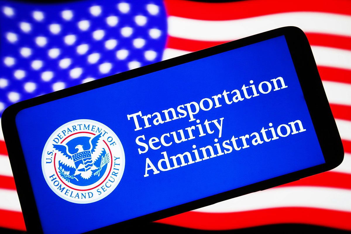 Transportation Security Administration (TSA) logo is seen on a smartphone and United States flag on a pc screen.